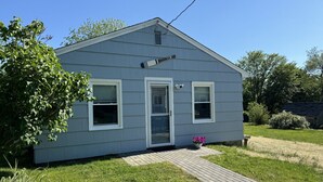 A single-story house with a grey exterior, a front door, and two windows. - The Oar Cottage (24) · walk Mystic-Train/EV Lvl-2 (Stonington)