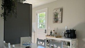 A dining area with a white table and chairs, a window with a view of greenery, a coffee maker on a white table, and a door leading outside. - The Oar Cottage (24) · walk Mystic-Train/EV Lvl-2 (Stonington)