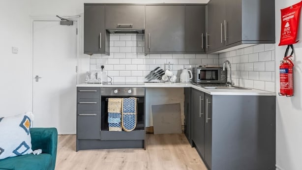 Une cuisine compacte avec des meubles gris, un four à micro-ondes, une bouilloire et un petit four. Un extincteur est fixé au mur et un canapé de couleur turquoise se trouve dans la pièce adjacente.