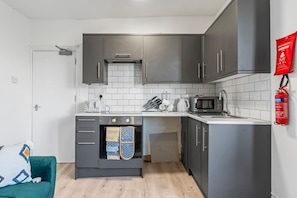 Une cuisine compacte avec des meubles gris, un four à micro-ondes, une bouilloire et un petit four. Un extincteur est fixé au mur et un canapé de couleur turquoise se trouve dans la pièce adjacente.