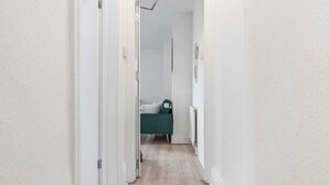 Un couloir étroit aux murs blancs et au plancher en bois mène à une pièce où se trouve un lit.