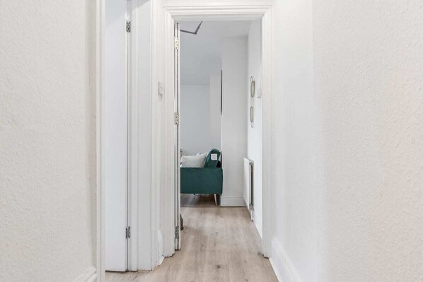 Un couloir étroit aux murs blancs et au plancher en bois mène à une pièce où se trouve un lit.