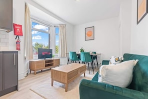 Un salon moderne avec un canapé vert, une table basse en bois et un téléviseur posé sur un meuble en bois. On y trouve également un coin repas avec des chaises de couleur turquoise et une table.
