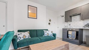 A compact living space with a green sofa, a wooden coffee table, and a kitchen area with dark cabinets and a tiled backsplash. - Cedar View – 2 Bed Apartment (Newport)