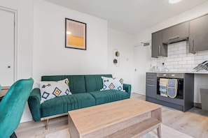 A compact living space with a green sofa, a wooden coffee table, and a kitchen area with dark cabinets and a tiled backsplash.