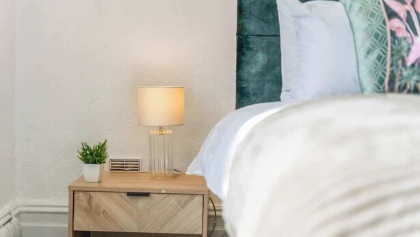A neatly made bed with a white comforter and pillows, a bedside lamp, and a small potted plant on a wooden nightstand. - Cedar View – 2 Bed Apartment (Newport)