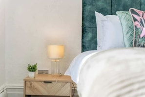 A neatly made bed with a white comforter and pillows, a bedside lamp, and a small potted plant on a wooden nightstand.