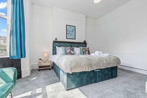 Un dormitorio con una cama, una mesita de noche, una silla azul y una ventana con cortina.
