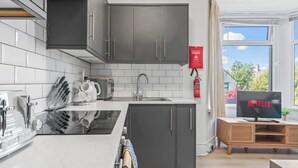 Une cuisine moderne avec des meubles de couleur sombre, un plan de travail blanc et une cuisinière encastrée. Il y a une fenêtre donnant sur des arbres et un extincteur rouge fixé au mur.