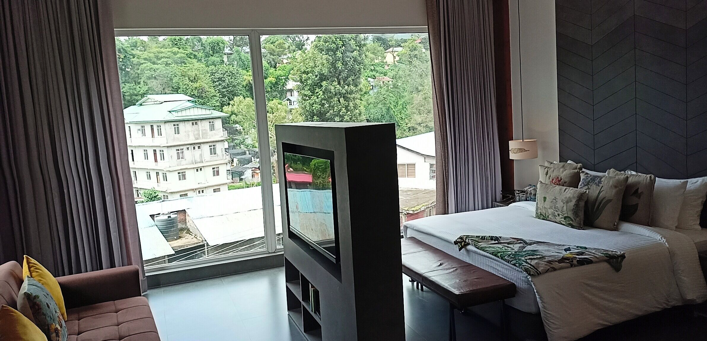 Ein Hotelzimmer mit großem Fenster, einem Bett, einem Fernseher und einem Sofa.