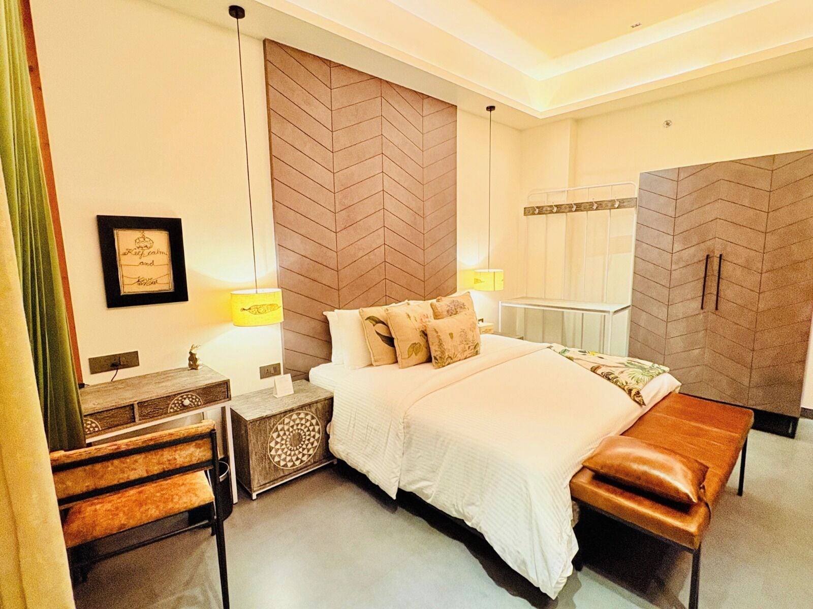 A hotel room with a bed, bedside table, lamp, bench, and a decorative wall panel.