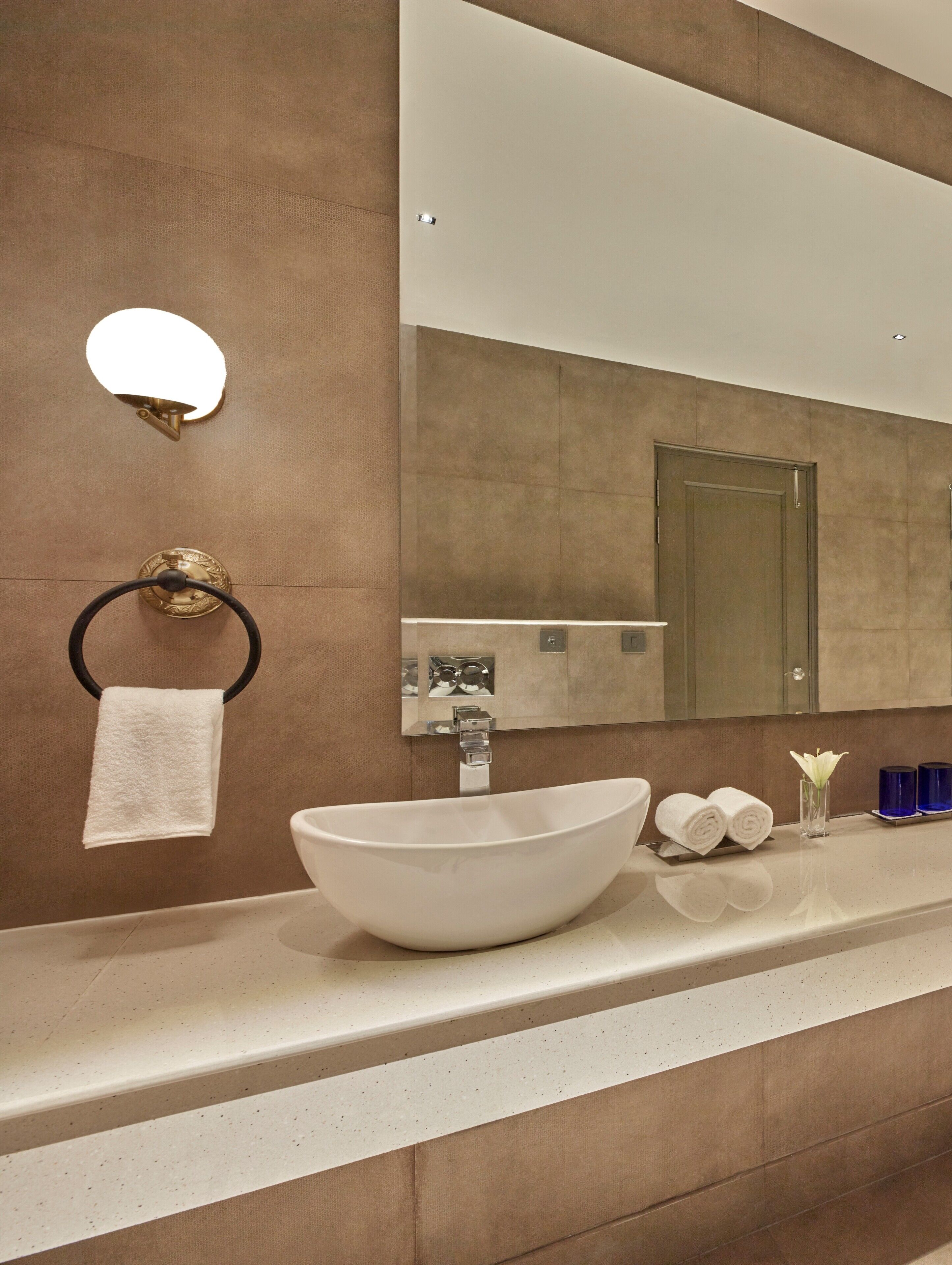 A modern bathroom with a large vessel sink, a towel ring, a mirror, and a small vase with a flower.