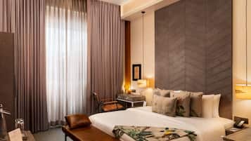 A hotel room with a large bed, a desk with a chair, a bench, and a large window with curtains.
