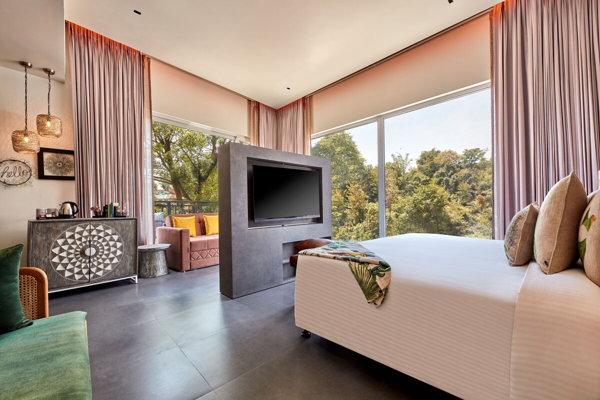 A modern bedroom with a large bed, a flat-screen TV, a sofa, and a view of trees through large windows.