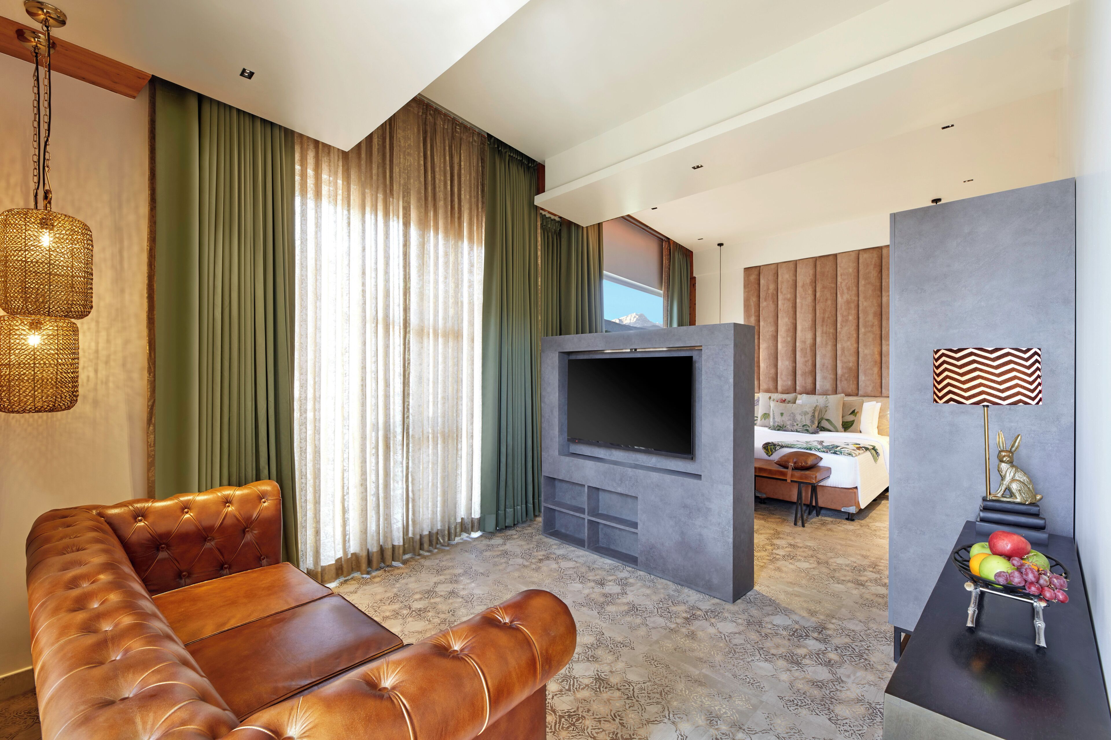 A modern hotel room with a leather sofa, a flat-screen TV, a dining area with fruit, and a view of the mountains.