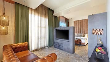 A modern hotel room with a leather sofa, a flat-screen TV, a dining area with fruit, and a view of the mountains.