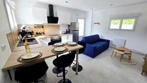 A modern kitchen with white cabinets, a black range hood, and a stainless steel refrigerator. There is a dining table with four chairs and a blue sofa in the living area.