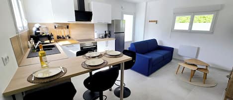 Eine moderne Küche mit weißen Schränken, einer schwarzen Dunstabzugshaube und einem Edelstahl-Kühlschrank. Im Wohnbereich befindet sich ein Esstisch mit vier Stühlen und ein blaues Sofa.