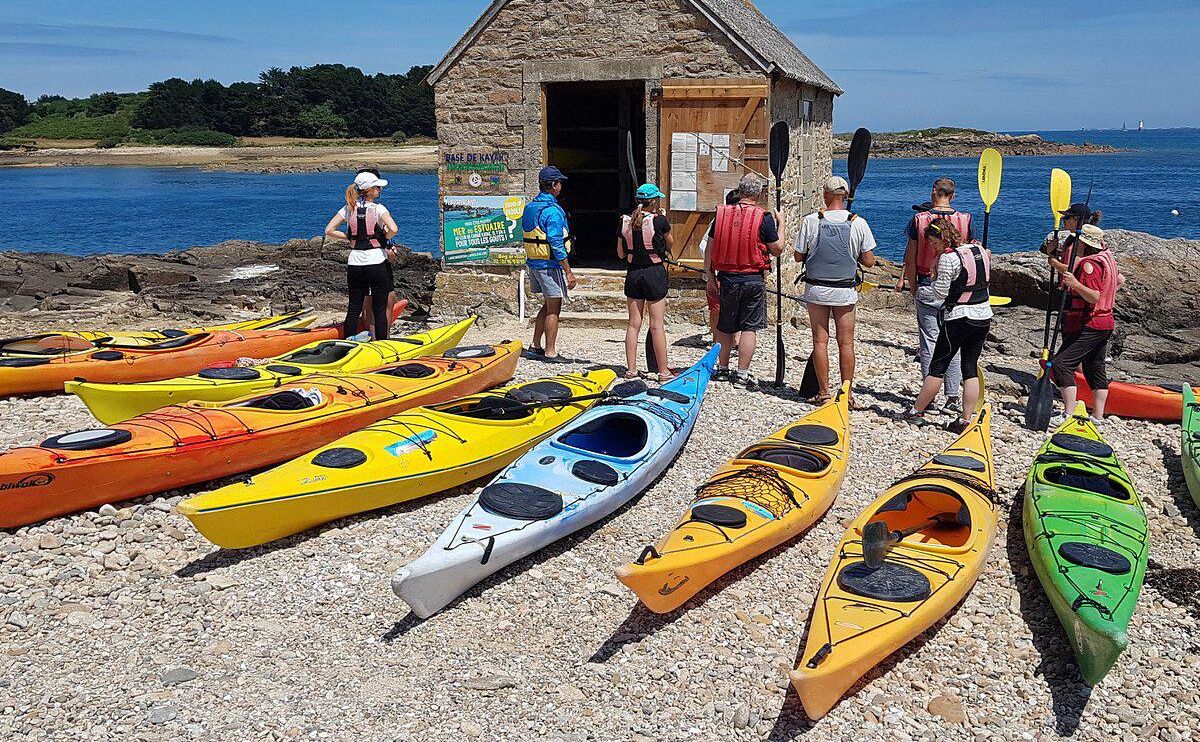 Un groupe de personnes se tient près de kayaks aux couleurs vives sur une plage de galets, avec un petit bâtiment en pierre en arrière-plan.