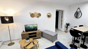 A modern living room with a flat-screen TV, a standing lamp, a dining area with a table and chairs, and decorative wall art.