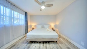 Ein Schlafzimmer mit einem Bett, zwei Nachttischlampen, einem Deckenventilator und großen Fenstern mit durchsichtigen Vorhängen.