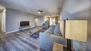 Ein geräumiges Wohnzimmer mit einem Flachbildfernseher, grauen Sofas, einem Couchtisch und einem Deckenventilator.