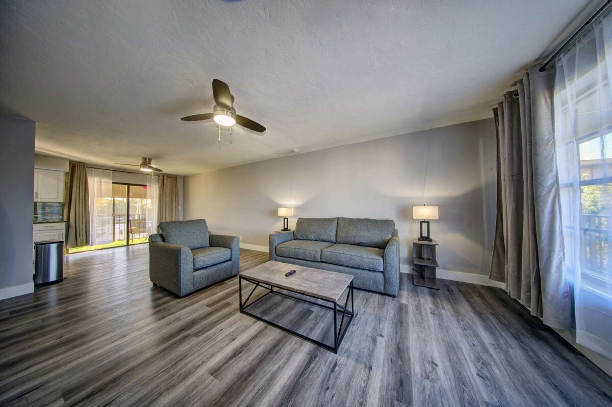 Ein modernes Wohnzimmer mit einem grauen Sofa, einem grauen Sessel und einem Couchtisch. Zwei Stehlampen und ein Deckenventilator sind vorhanden. Das Zimmer verfügt über ein großes Fenster mit Vorhängen und Blick ins Freie.
