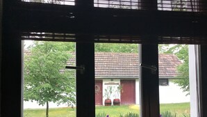 Udsigt gennem et vindue med en dør, der viser en bygning med en rød dør og et tegltag, omgivet af grøn vegetation og lilla blomster.