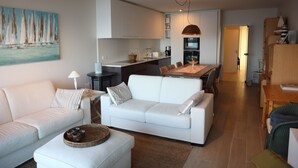 Una sala de estar moderna con un sofá blanco, un comedor con una mesa de madera y una cocina al fondo.