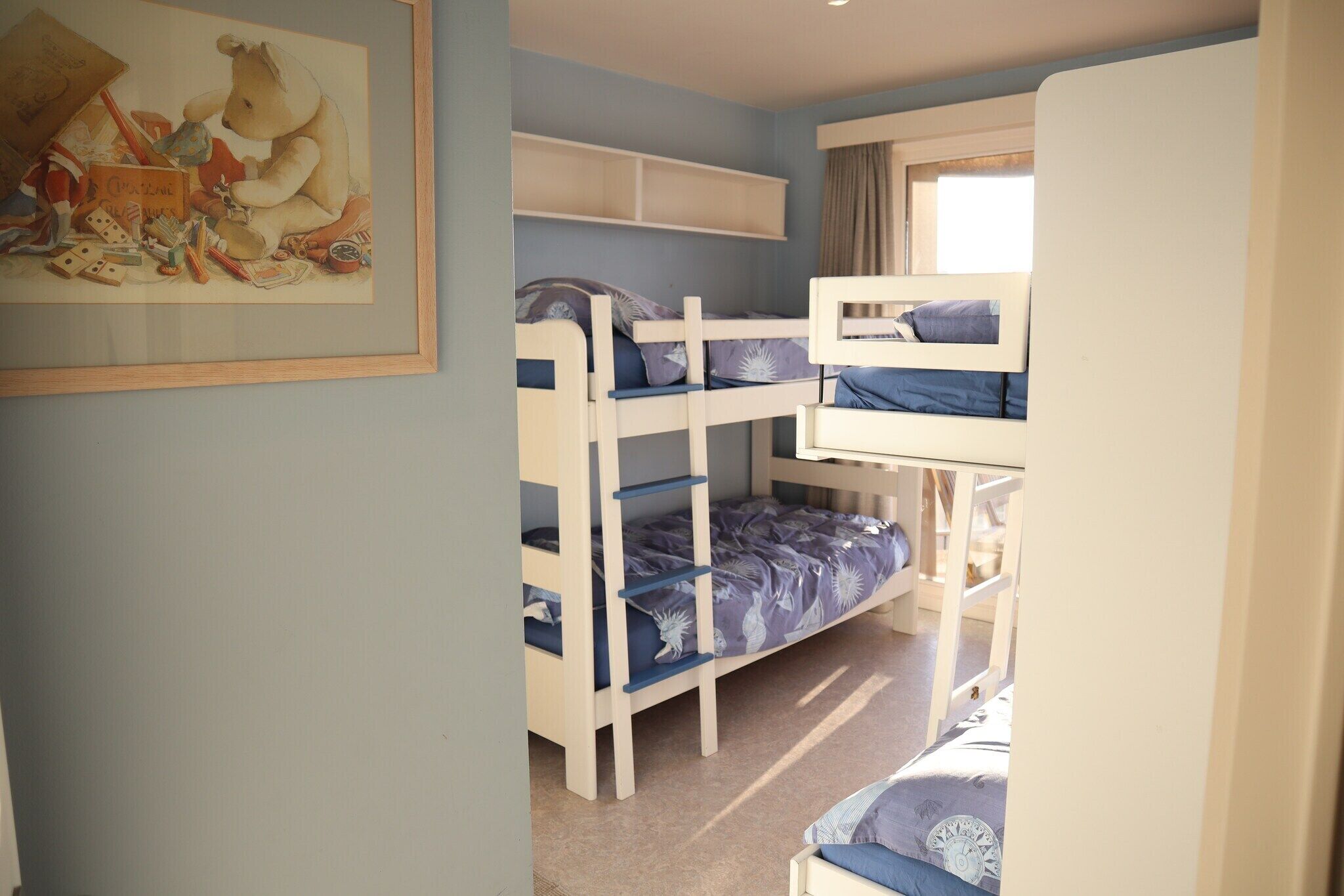 A bunk bed room with a framed picture on the wall, a window with curtains, and a small table.