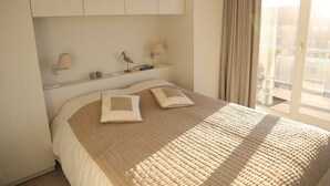 Una cama bien hecha con colcha color beige, dos almohadas y una mesita de noche con lámpara y un objeto decorativo.