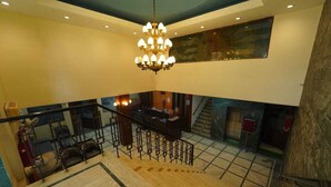 A spacious interior with a high ceiling, a central chandelier, a staircase, and a reception desk. - Hotel Saket (Prayagraj)