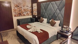 A hotel room with a large bed, a grey upholstered sofa, a wooden nightstand with a coffee maker, and a wall with floral artwork. - Hotel Saket (Prayagraj)