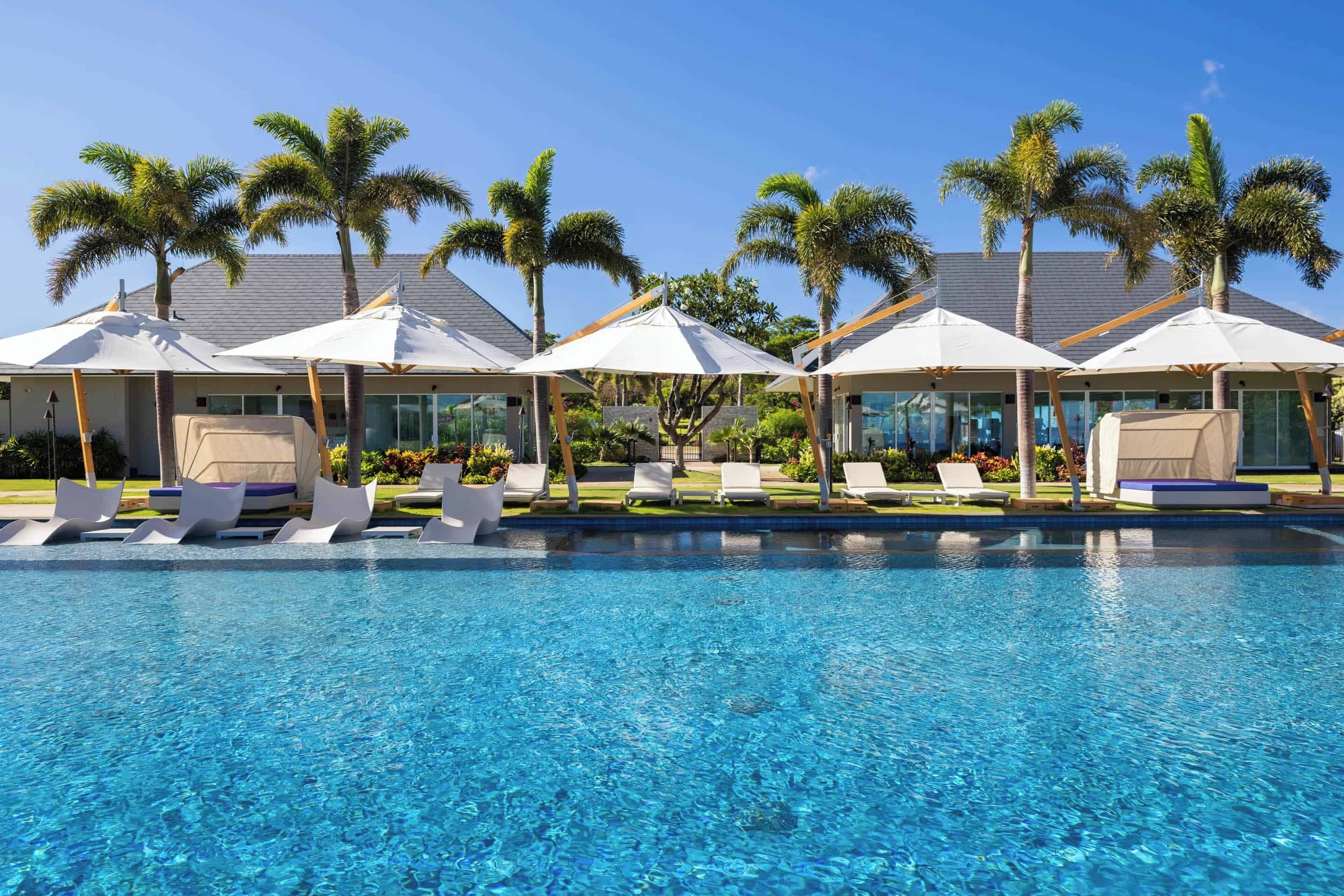 A swimming pool with lounge chairs and umbrellas, surrounded by palm trees and a building with a tiled roof.