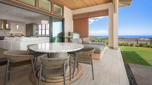A modern outdoor living area with a round table and wicker chairs, a built-in kitchen, and a view of the ocean. - Amaui Villas 11 Three Bedroom (Kamuela)