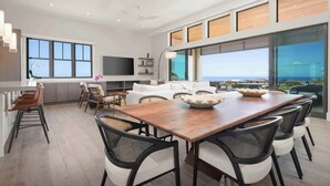 A spacious living area with a wooden dining table, white sofas, and black chairs. There is a large window offering a view of the ocean. - Amaui Villas 11 Three Bedroom (Kamuela)