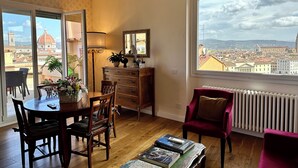 A room with a dining table, chairs, a red armchair, a wooden dresser, a mirror, and a view of a cityscape with a dome and buildings. - Florence Rooftop Apt - Jacuzzi & Panoramic View (Firenze)
