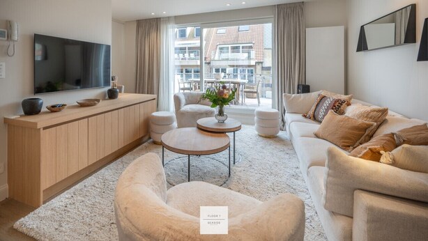 A modern living room with a sofa, coffee table, and TV. There is a large window with curtains, a wooden cabinet, and a small table with a vase.