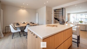 A modern kitchen with a central island, dining area with chairs, and a staircase leading to another level.