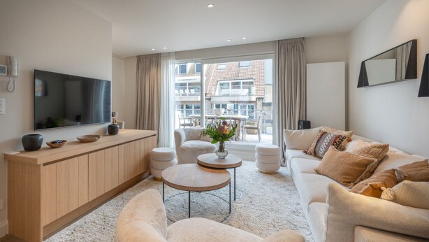 Una sala de estar moderna con sofá, mesa de centro y televisor. Hay un ventanal con cortinas, un mueble de madera y una pequeña mesa con un florero.