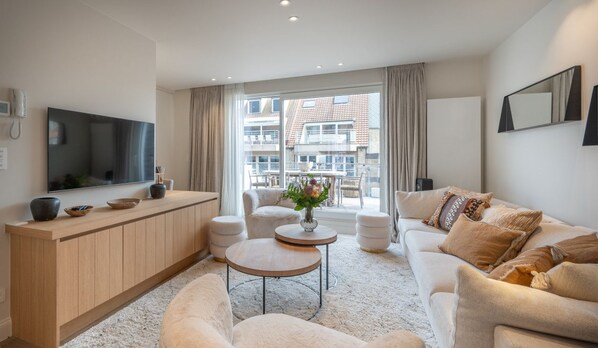 A modern living room with a sofa, coffee table, and TV. There is a large window with curtains, a wooden cabinet, and a small table with a vase.