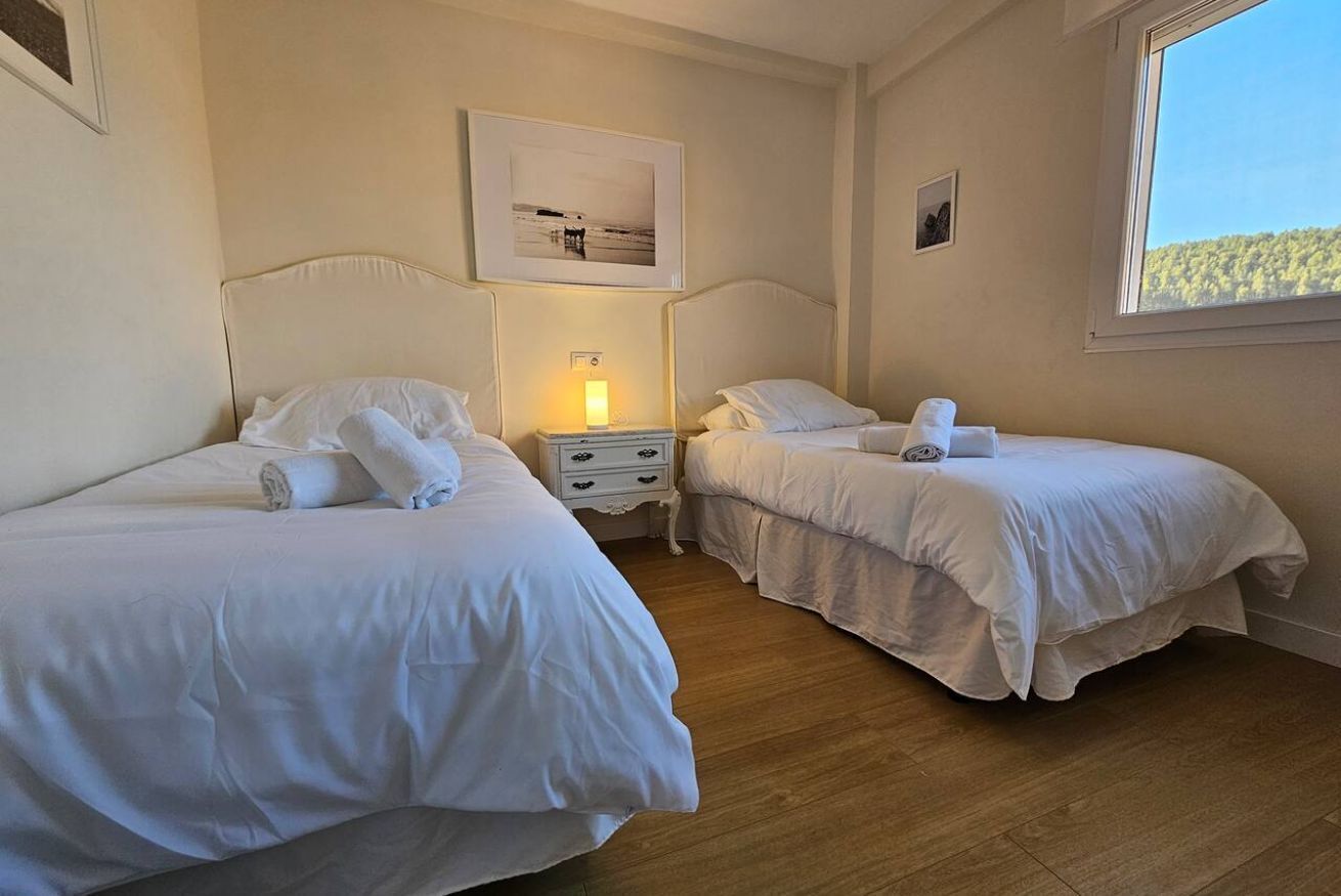 A room with two beds, a nightstand with a lamp, and a window with a view of trees.