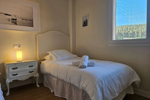 Een net opgemaakt bed met witte lakens, een bedlampje en een lijst met een foto aan de muur.
