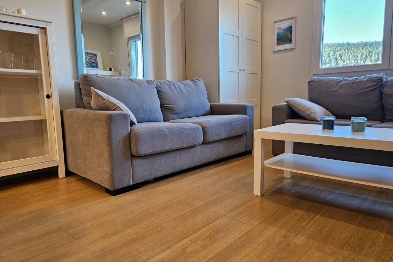 Un salon moderne avec un canapé gris, une table basse blanche et une vue sur une forêt par la fenêtre.