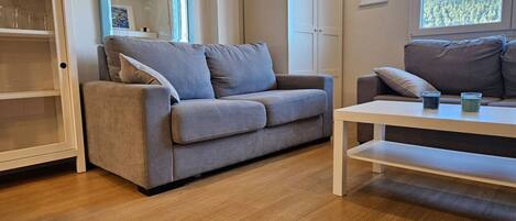 A modern living room with a grey sofa, a white coffee table, and a view of a forest through a window.