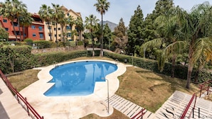 A swimming pool with a curved edge, surrounded by palm trees and a hedge, with a multi-story building in the background. - Amazing Sea and Golf Course view 2BR, Parking Pool REF037 (Benalmádena)