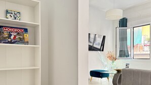 Una sala de estar moderna con un sofá gris, un sillón azul y una mesa de centro de vidrio. Hay un juego de Monopoly en una estantería, y un televisor montado en la pared.