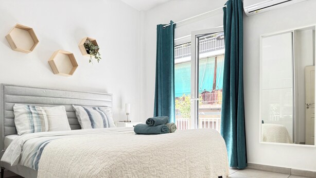 Un dormitorio con cama, mesita de noche, estante en la pared con una planta y vistas a un balcón a través de una ventana con cortinas.