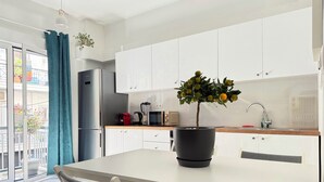 Cocina moderna con gabinetes blancos, un refrigerador de acero inoxidable, un microondas y un fregadero. Hay una mesa con dos sillas y una planta en maceta encima.