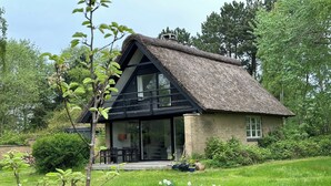 A thatched-roof cottage with a glass door entrance, a balcony, and a garden with white flowers. - Cozy, thatched cottage with modern décor (Knebel)